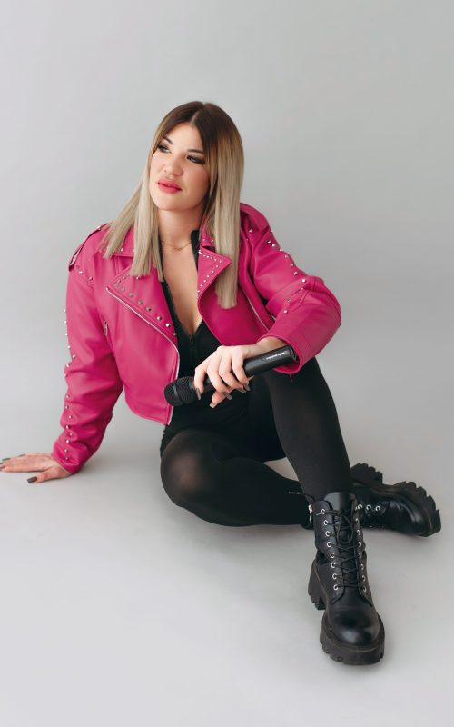 KLAU énekesnő és retro DJ mikrofonnal, földön ülve, fekete ruhában, pink kabátban, világos stúdióban