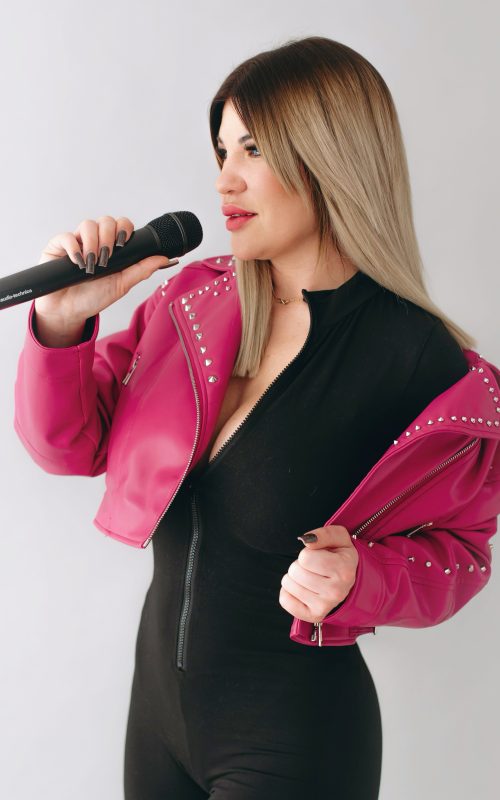 KLAU énekesnő és retro DJ mikrofonnal, fekete ruhában, pink kabátban, világos stúdióban
