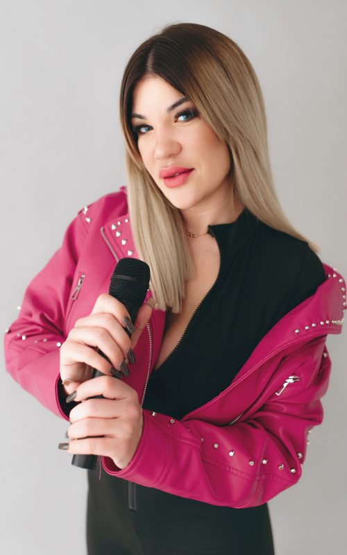 KLAU énekesnő és retro DJ mikrofonnal, fekete ruhában, pink kabátban, világos stúdióban