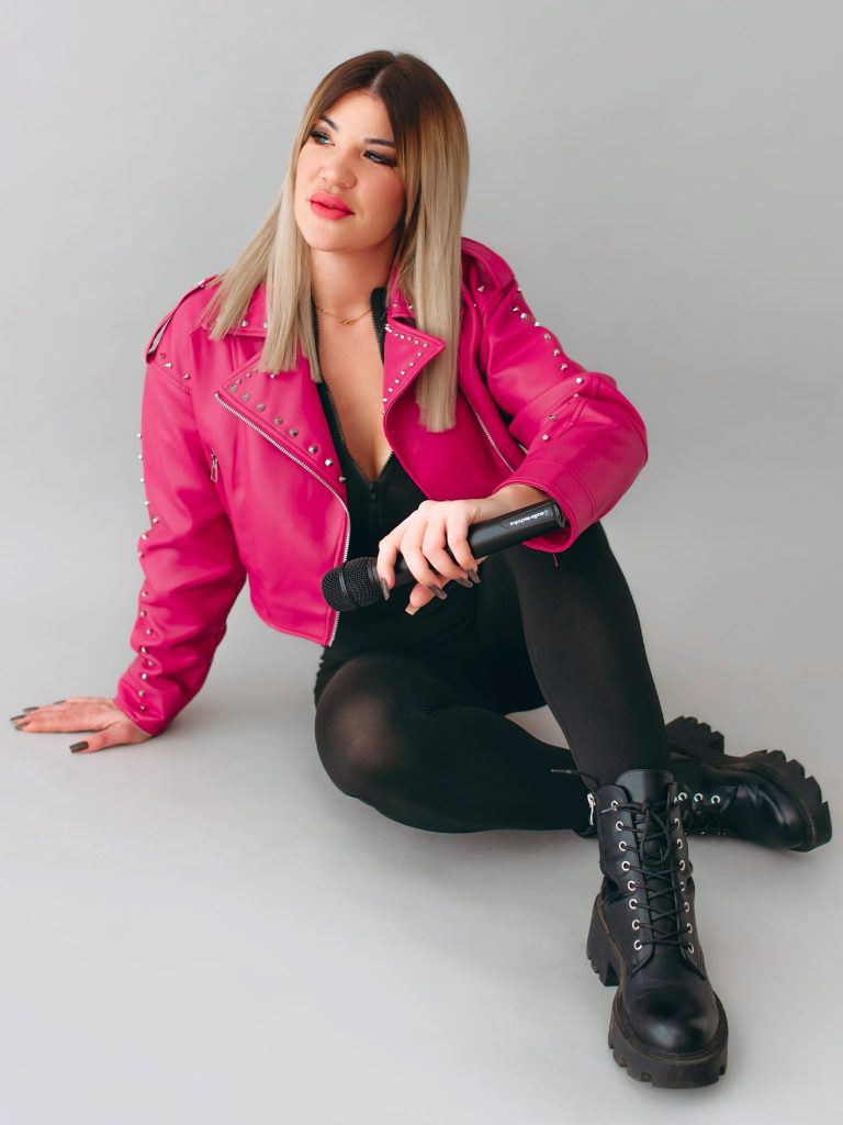 KLAU énekesnő és retro DJ mikrofonnal, fekete overálban, pink kabátban, világos stúdióban
